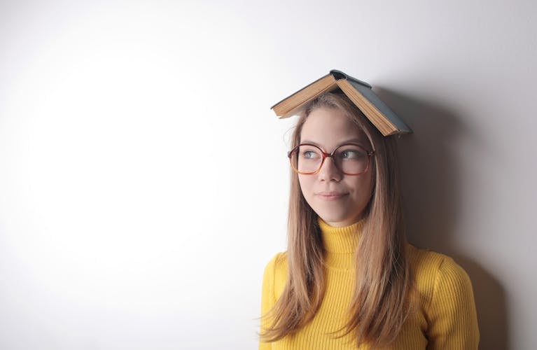A woman wearing glasses with a book on her head, playfully posing in a yellow sweater.